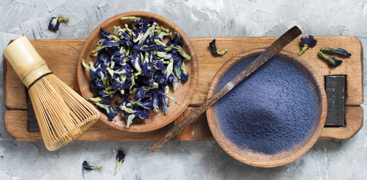 bowl of butterfly pea blue matcha powder with whisk and dried flowers